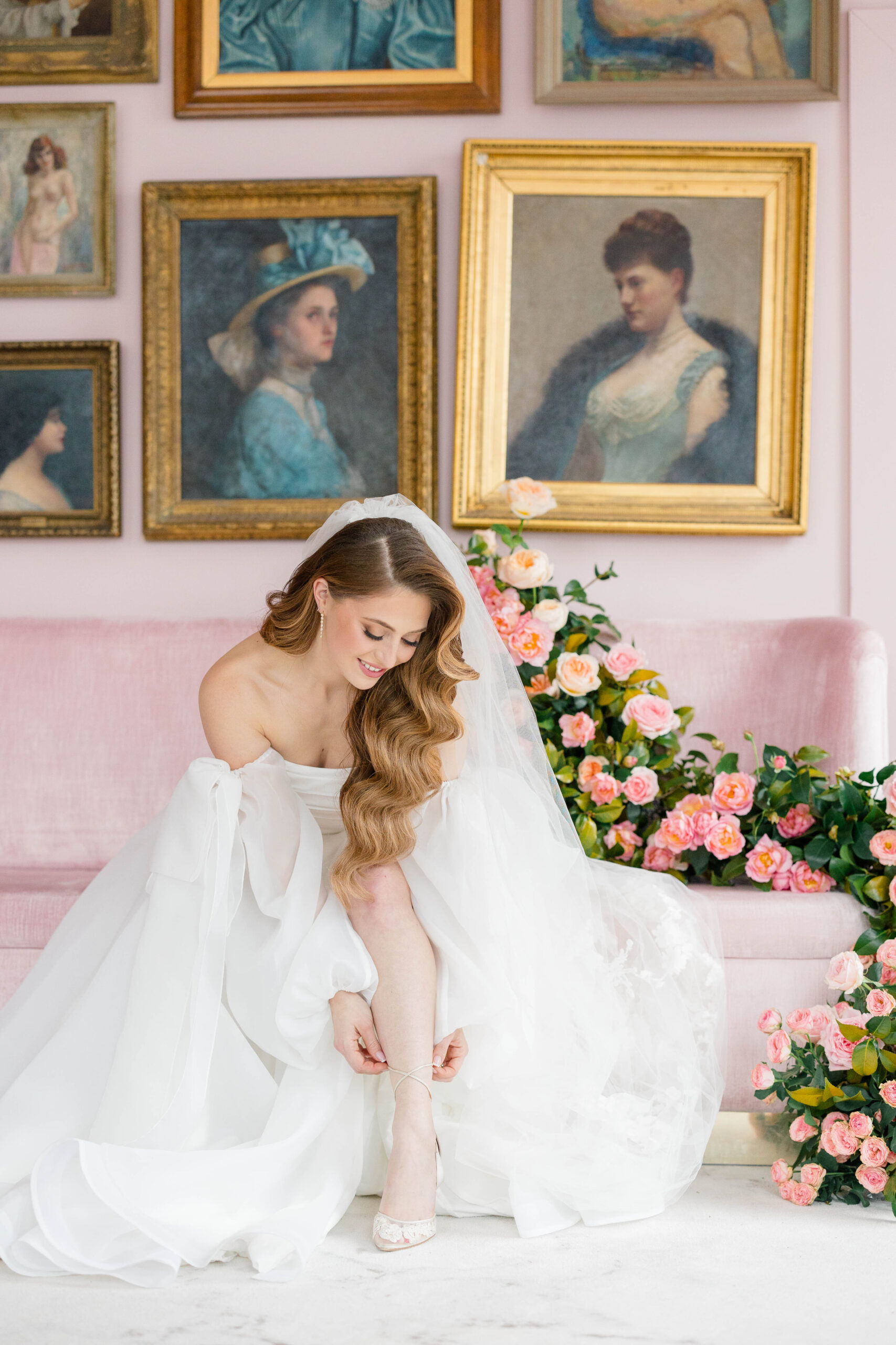 Newport Wedding Photographer and Videographer | Serving CT, RI, Massachusetts, and Surrounding New England Areas | Bride fixing her shoe on a pink couch with paintings on the wall